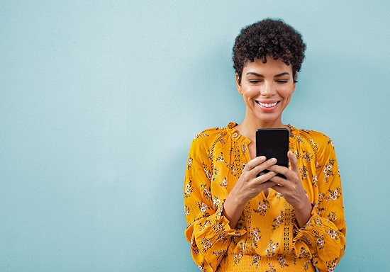 Mujer afroamericana sonriendo, con un teléfono inteligente y de pie contra una pared azul claro.