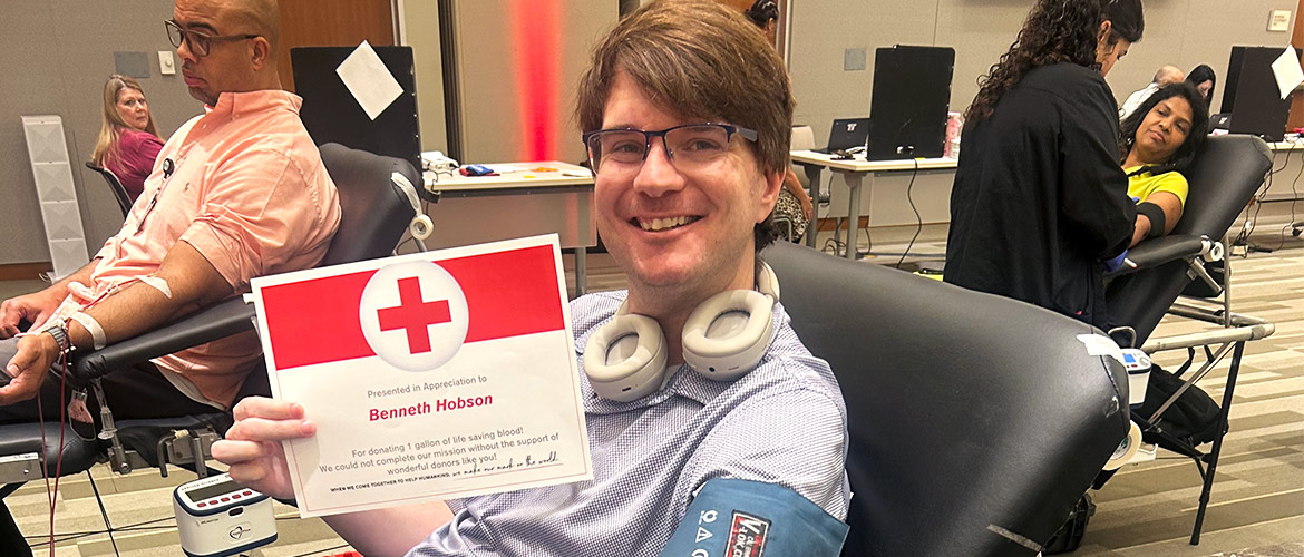 A smiling man wearing glasses sits in a reclining chair to donate blood and holds a certificate from the American Red Cross