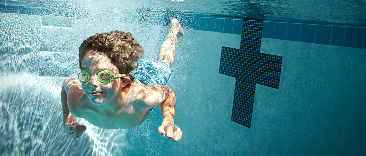 Boy with goggles swims underwater in the deep end of a pool. 