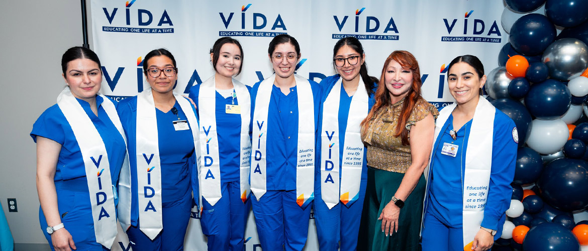 Seven women stand in front of a backdrop with the VIDA logo and the words "Educating One Life at a Time." 