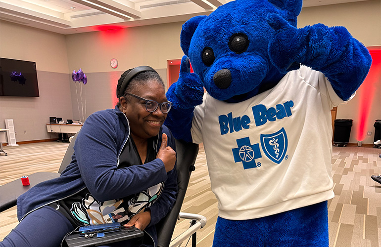 A woman in a blue track suit and glasses gives a thumbs up and smiles for a photo with Blue Bear, the Blue Cross and Blue Shield of Texas mascot. 
