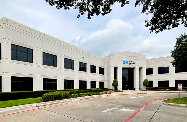 A two-story white office building with a Blue Cross and Blue Shield of Texas sign above the front entrance and a driveway and green landscaping in the foreground. 