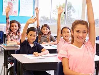Middle school-aged children seated at their desks in a classroom with their hands raised