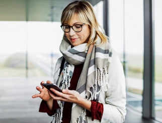 Mujer caucásica rubia con suéter, bufanda y lentes mirando su teléfono celular.