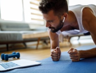 Hombre joven haciendo un ejercicio de plancha en la alfombra de su sala de estar, viendo un video de ejercicios o una transmisión en vivo en su computadora portátil.
