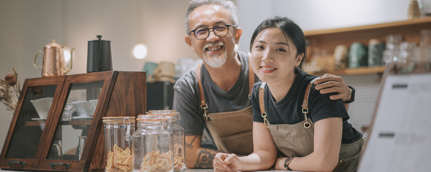 An older man and young woman wearing aprons smile behind a shop counter