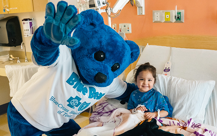 La mascota Blue Bear de BCBSTX visita a una niña pequeña en la cama de un hospital.