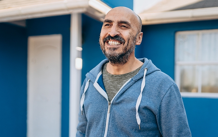 Hombre de barba y con sudadera con capucha sonríe delante de una casa pintada de azul.