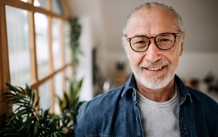 Older man in glasses smiles near plants bordering a window. 