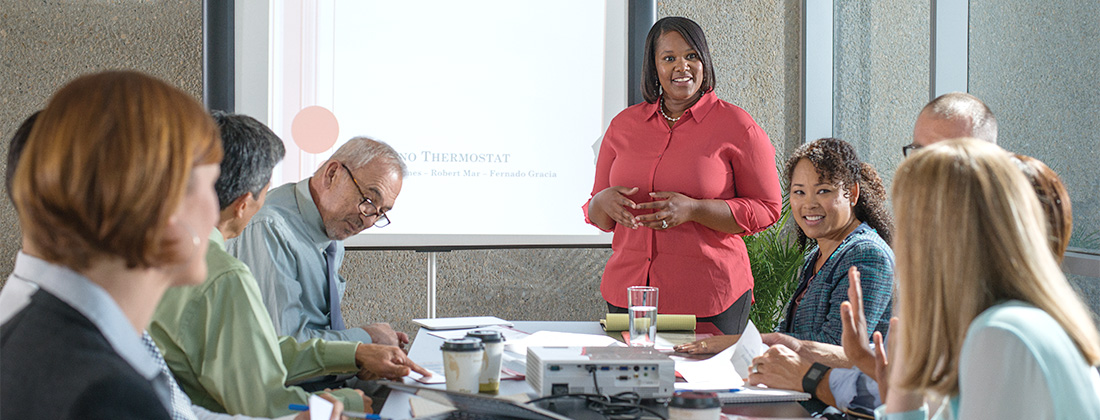 Multi-generational employees seated around desk listen to a presentation. 